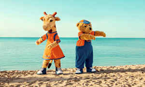 To maskoter, en giraff og en bjørn, står på en strand med havet i bakgrunnen. (AI-generert tekst)