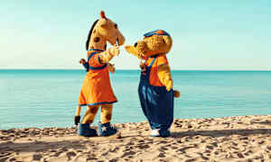 To maskoter, en giraff og en bjørn, står på en sandstrand med havet i bakgrunnen. (AI-generert tekst)