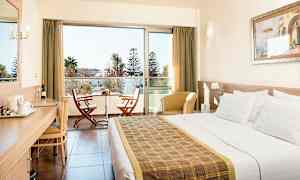 Et hotellrom med en dobbeltseng, skrivebord og stoler ved en balkong. (AI-generert tekst)