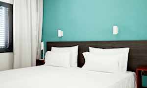 Hotellrom med en oppredd seng med hvitt sengetøy og blå vegg. (AI-generert tekst)