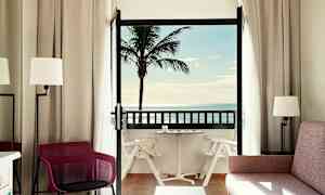 Et hotellrom med balkong som viser en palme og havutsikt. (AI-generert tekst)