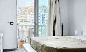 Utsikt fra et hotellrom, med seng og en åpen balkongdør. Balkong med stoler og utsikt over bygninger. (AI-generert tekst)