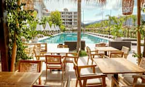 Bilde av et hotellområde med solsenger ved siden av et rektangulært basseng og trebord i forgrunnen. (AI-generert tekst)