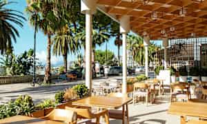 Utendørs servering med trebord og stoler, omgitt av palmer og bymiljø. (AI-generert tekst)