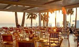 Utendørs servering med bord og stoler ved solnedgangen, med palmer og havet i bakgrunnen. (AI-generert tekst)