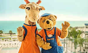 To maskoter, en sjiraff og en bjørn, står foran havet i bakgrunnen. (AI-generert tekst)