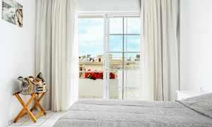 Et lyst soverom med en grå oppredd seng, en trestol med en sammenrullet veske, gardiner og en balkongdør som leder til en utsikt med blomster og bygninger. (AI-generert tekst)