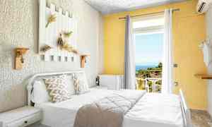 Lyst hotellrom med seng, dekorerte puter og utsikt mot balkong med hav og grøntområder. (AI-generert tekst)