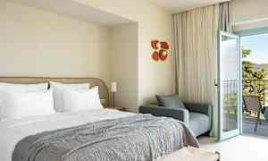 Hotellrom med oppredd seng, lenestol og åpen balkongdør. (AI-generert tekst)