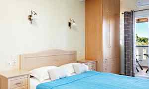 Et soverom med en dobbeltseng som har et blått sengeteppe og to puter, et garderobeskap og nattbord. En utendørs balkong og gardiner er synlige i bakgrunnen. (AI-generert tekst)