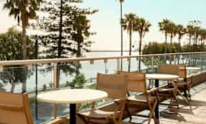 Tre stoler og bord på en balkong med utsikt over palmer og hav. (AI-generert tekst)