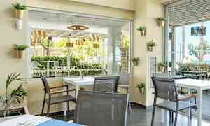 En moderne innredet kafé med bord og stoler, utsikt mot grønne trær og utemøbler. (AI-generert tekst)