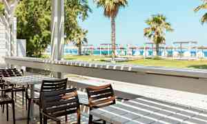 En terrasse med bord og stoler, med utsikt over en grønn gressplen, palmer og stranden i bakgrunnen. (AI-generert tekst)