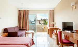 Et hotellrom med to senger, en sofa, et bord, et skrivebord med stol og en balkong med utsikt. (AI-generert tekst)