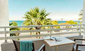 En balkong med utsikt over en palme, havet og nabobygninger. (AI-generert tekst)