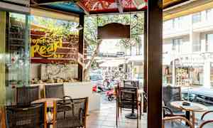 Et caféområde med sitteplasser og bord ved siden av et vindu. Utenfor sees en gate med butikker og biler. (AI-generert tekst)