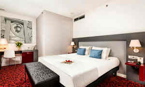 Et hotellrom med en stor seng, to blå puter, et skrivebord med stol, rød teppe og moderne lamper. (AI-generert tekst)