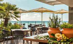 En utendørs terrasse med bord og stoler under parasoller, med havet i bakgrunnen og grønne planter samt eksotiske frukter på et bord i forgrunnen. (AI-generert tekst)
