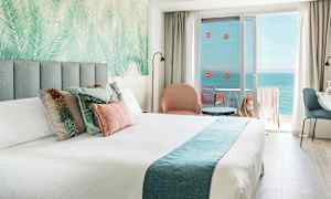 Et hotellrom med en dobbeltseng, dekorerte puter, en sittegruppe og en balkong med havutsikt. (AI-generert tekst)