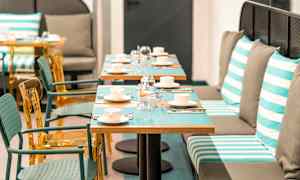 Bilde av en restaurant med bord dekket med kopper, glass og bestikk. Stoler og benkesitteplasser med stripete puter vises. (AI-generert tekst)