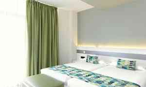 Et hotellrom med to enkeltsenger, grønne gardiner og matchende sengedekorasjoner. (AI-generert tekst)