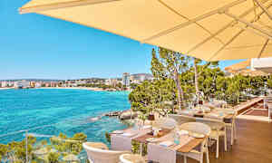 Uteservering med utsikt over en klarblå havsbukt og bygninger i det fjerne. (AI-generert tekst)