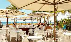 Utendørs restaurant med hvite bord og stoler under parasoller, utsikt mot havet. (AI-generert tekst)