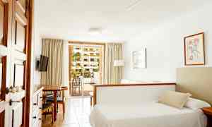 Et hotellrom med en seng, sofa og spisebord, samt utsikt mot en balkong med grønne planter utenfor. (AI-generert tekst)