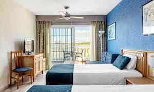 Bilde av et hotellrom med to senger, en blå vegg og en glassdør mot en balkong. (AI-generert tekst)