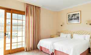 Et hotellrom med to enkeltsenger med hvitt sengetøy, et bilde på veggen og en glassdør med gardiner. (AI-generert tekst)