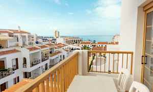 Utsikt fra en balkong med et bord og stoler over hvite bygninger med røde tak og havet i bakgrunnen. (AI-generert tekst)