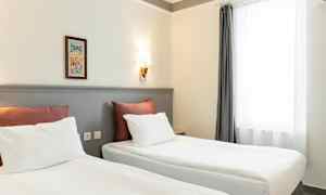 To senger med hvitt sengetøy og røde puter står i et rom med lyse vegger og en vegglampe. Et vindu med hvite gardiner er til høyre. (AI-generert tekst)