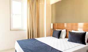 Et lyst hotellrom med en dobbeltseng, to mørke puter og et vindu med gardiner. (AI-generert tekst)