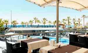 Utsikt fra en utendørs servering mot et bassengområde med parasoller og palmer samt havet i bakgrunnen. (AI-generert tekst)