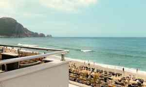 Et bilde av en strand med solsenger, parasoller og mennesker som svømmer i havet. I bakgrunnen vises en fjellklippe. (AI-generert tekst)