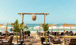 Utsikt over en strand med solsenger og parasoller, og en sti forbi palmeplanter leder mot havet. (AI-generert tekst)