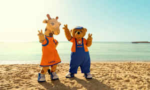 To maskotkarakterer med navnene Lollo og Bernie står på en strand foran det klare havet. (AI-generert tekst)