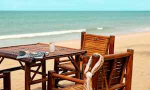 Stoler og bord av tre står på en strand med havet i bakgrunnen. (AI-generert tekst)