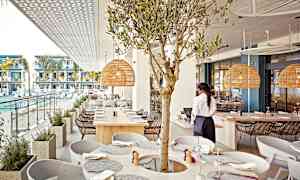 En restaurant med utsikt over et basseng og moderne leiligheter, inkluderer en oliventredekorasjon og rottinglamper. (AI-generert tekst)