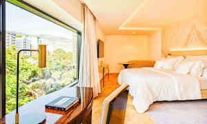 Et hotellrom med en stor seng og et skrivebord nær et stort vindu som viser utsikt over byen. (AI-generert tekst)