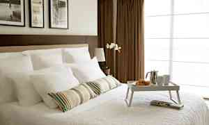 Et rom med en oppredd dobbeltseng, hvitt sengetøy og et frokostbrett med croissanter, en kaffekanne og kopper. I bakgrunnen sees brune gardiner og bilder på veggen. (AI-generert tekst)