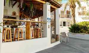 En utendørsbar med trestoler og tak av terrakottafliser. En palme og bygninger kan sees i bakgrunnen. (AI-generert tekst)