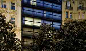 Bildet viser en moderne bygning med mørke glassfasader og klassiske bygninger på hver side, med en restaurant og parkering i forgrunnen. (AI-generert tekst)