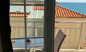 Utsikt fra et rom mot en balkong med bord og stoler, tak med røde takstein og havet i bakgrunnen. (AI-generert tekst)