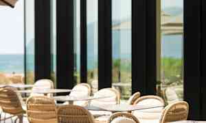 En uteplass med bord og stoler, ved siden av en bygning med store glassvinduer. (AI-generert tekst)