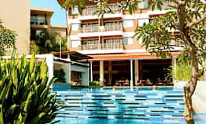 Et basseng med klart blått vann omgitt av planter og et hotell i bakgrunnen. (AI-generert tekst)