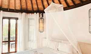 Interiør av et soverom med et tak laget av flettet tre, en hvit seng med myggnett og vinduer som slipper inn naturlig lys. (AI-generert tekst)