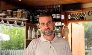 En hvit skjorte ved en bar med hyller fylt med flasker i bakgrunnen, samt et cocktailglass med en farget drikk foran seg. (AI-generert tekst)