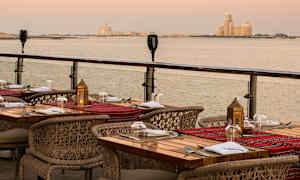 En utendørsrestaurant med dekkede bord og utsikt over havet ved solnedgang. (AI-generert tekst)