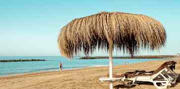 Bilde av en sandstrand med to solsenger under en halmparasoll og personer som går langs vannet. (AI-generert tekst)